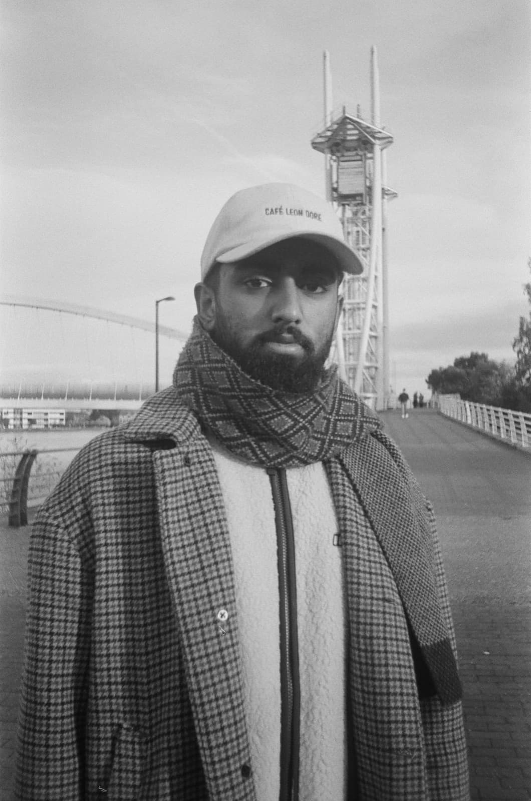 Aqeeb Munir, a black-and-white photograph on a footbridge, in a cap, patterned scarf and houndstooth coat
