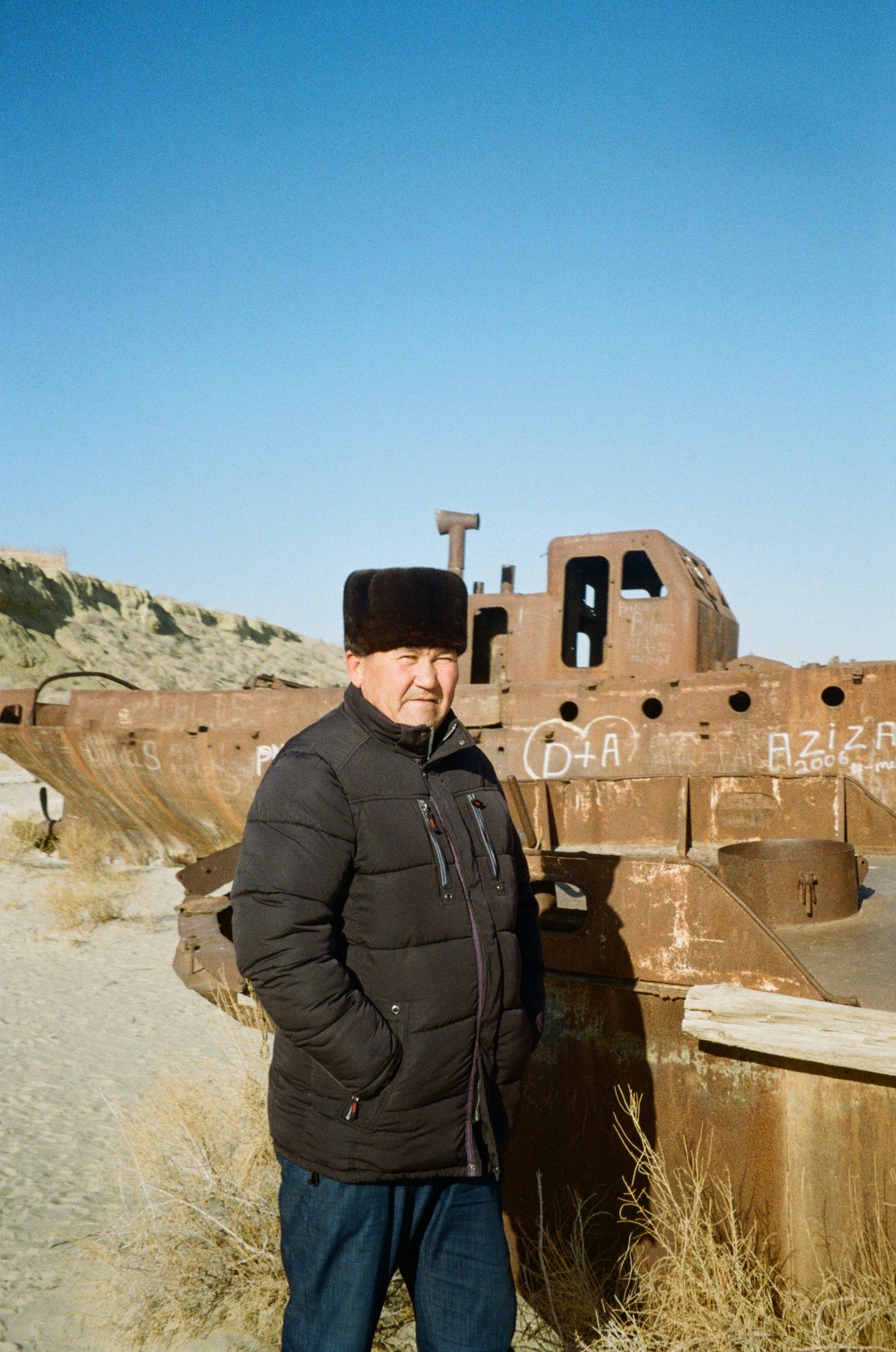 A man in a hat and dark coat standing before mud-brick ruins